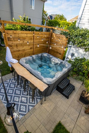 A Hydropool Serenity Hot Tub Is the focal point, it has a wooden live edge bar table and stools right next to it, with a wooden fence to the left and outdoor rug below, fiarylight hang above it. It is in the small garden of an airbnb. 