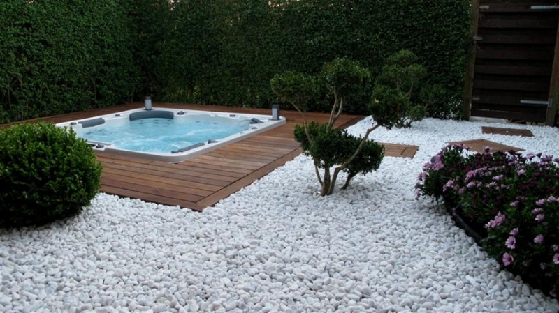 An inground hot tub surrounded by wooden deck, then white stones.