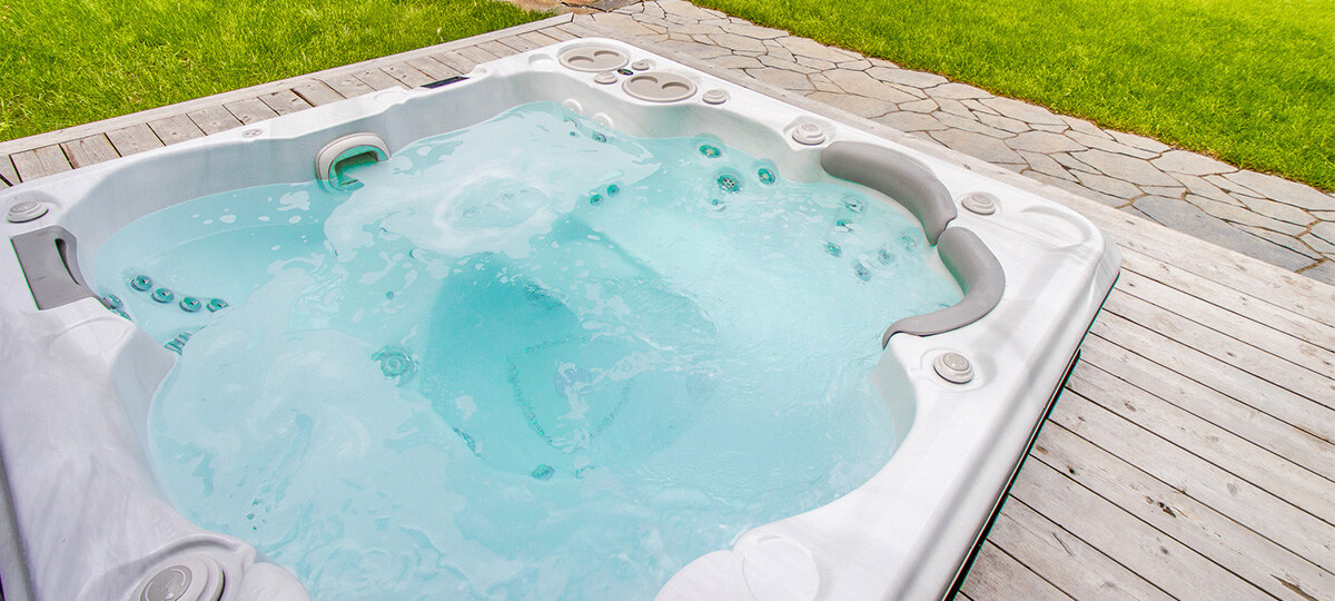 Bright blue water inside a white hot tub set inside a grey raw wood deck, with a lime green grass field in the background.