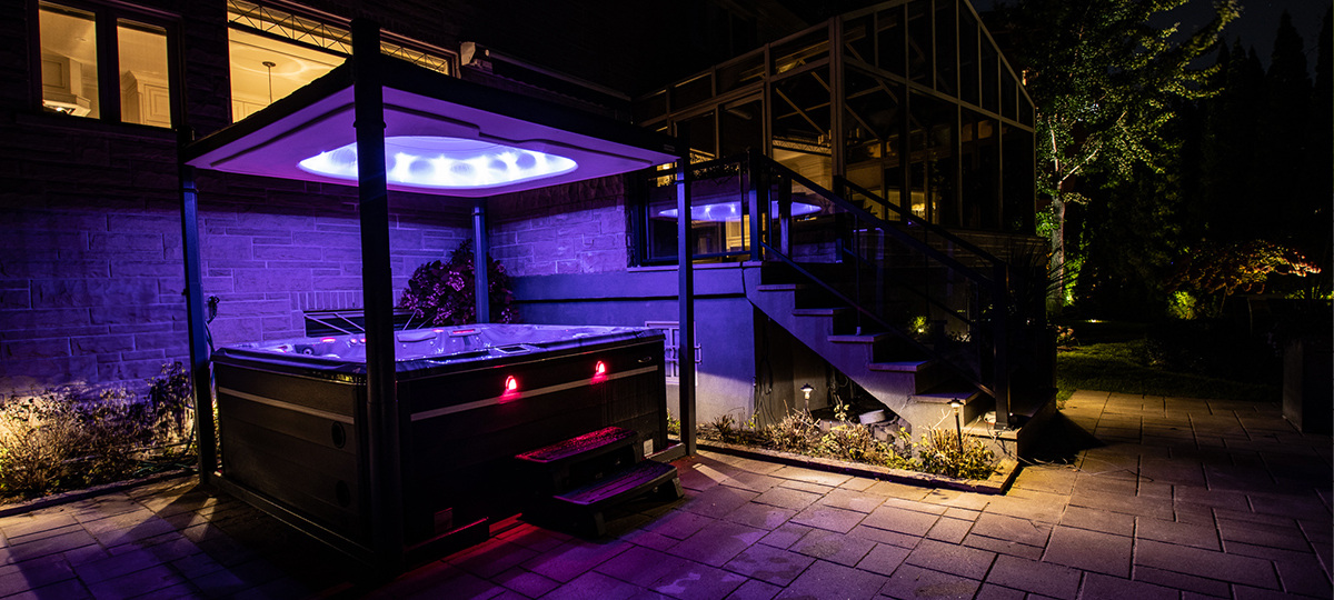 A Self Cleaning Hot Tub with two red lights on the side, covered by a covana electric hot tub cover lifted with purple lights shining from inside the cover soaking the entire area in purple light. The picture was taken at night, yellow lights shining from the background house and porch.
