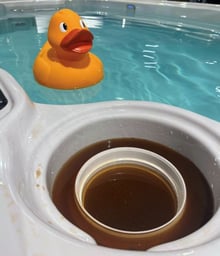 The filter compartment of a swim spa sitting in muddy water with clean blue swim spa water behind, with a yellow rubby ducky floating in the pool.