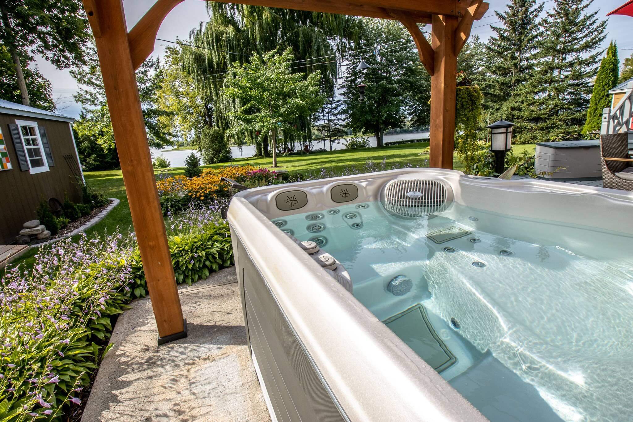 A high quality hot tub with a wooden pergola above it, surrounded by greenery, trees, and flowers.
