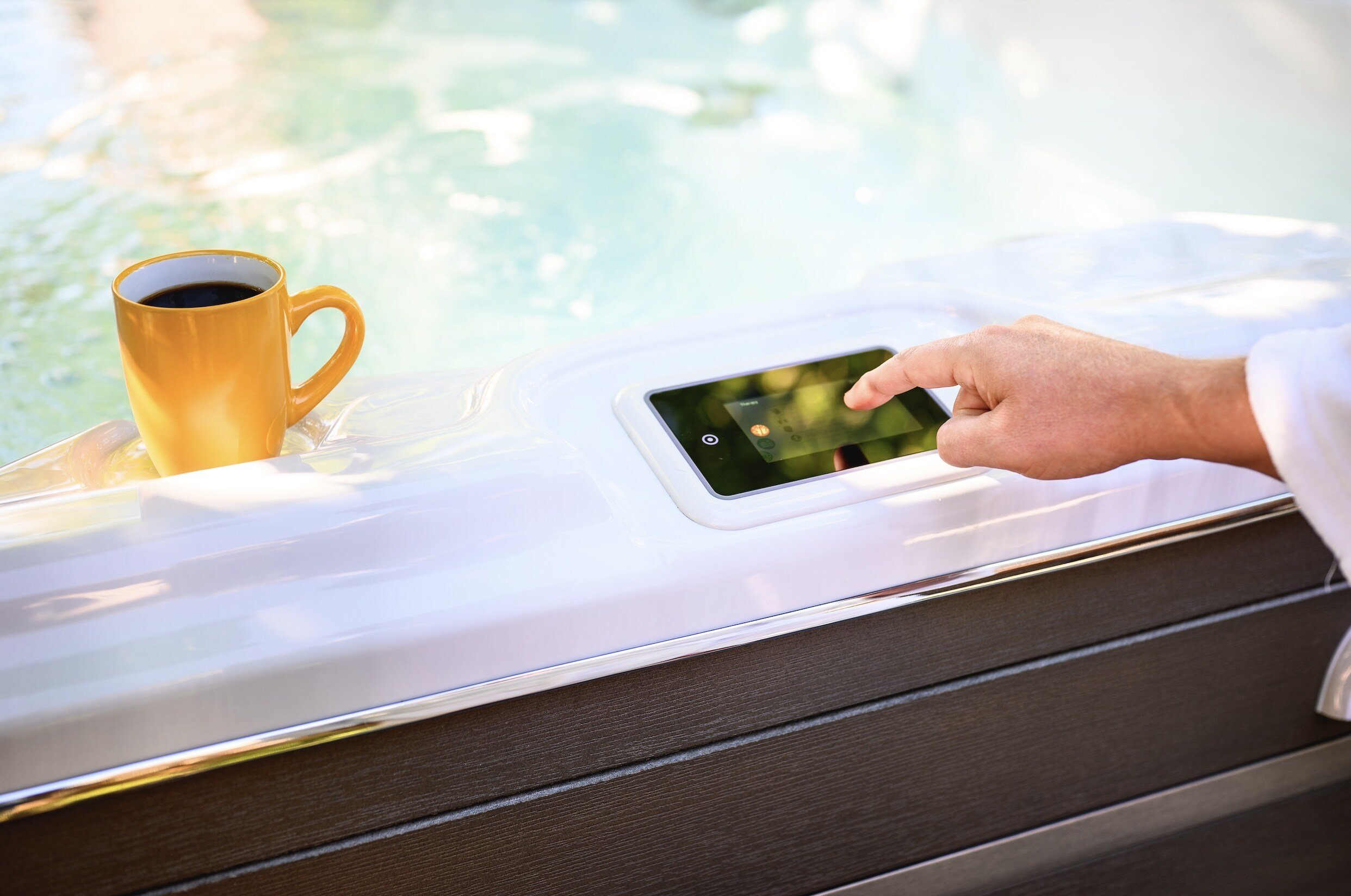 Man's hand pointing at the hydropool hot tub topside control, mustard yellow coffee cup resting on the side of the tub with cerulean hot tub water beyond. 