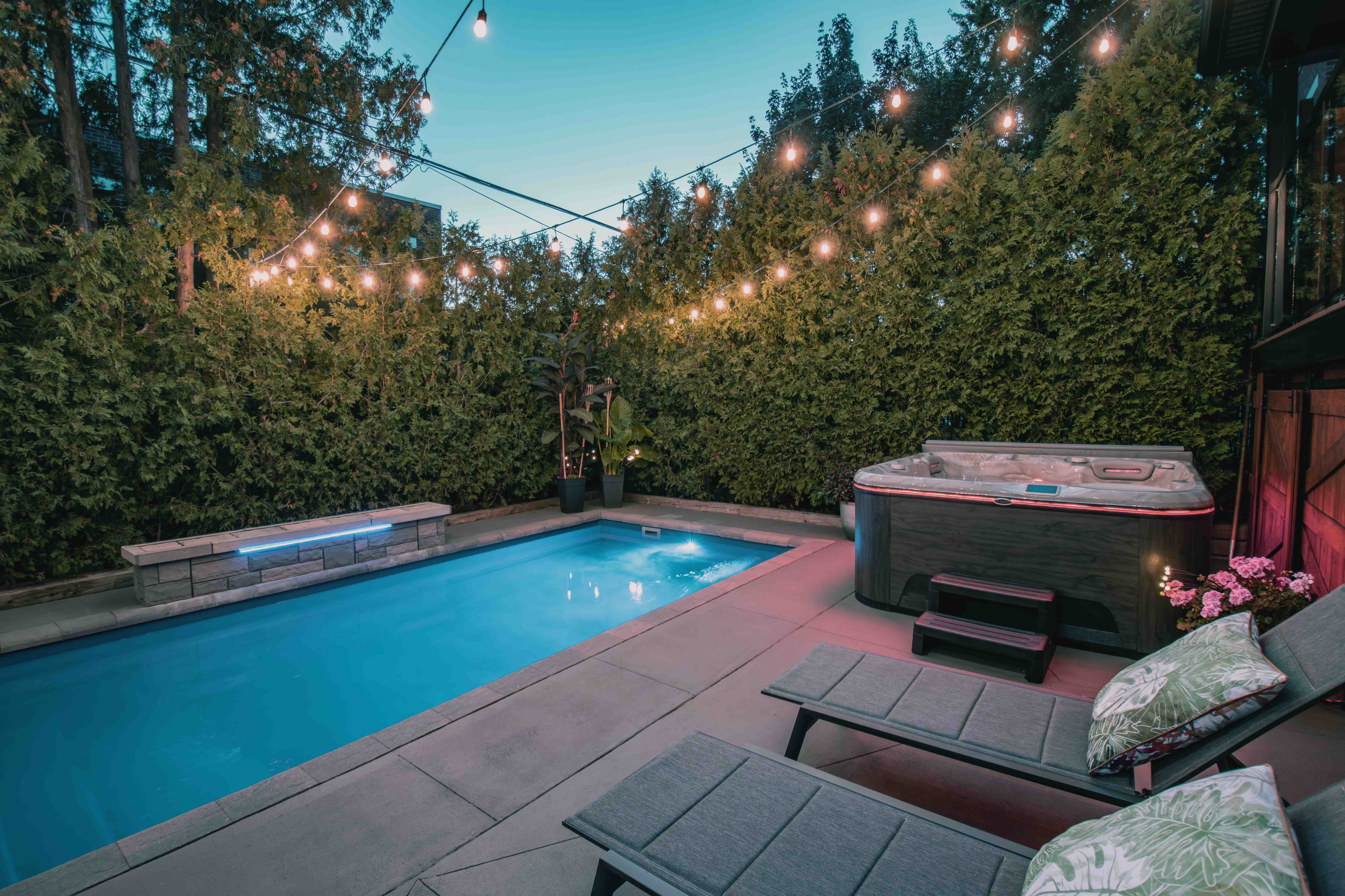 A styled garden with hung fairylights is surrounded by hedges. It includes a blue-watered built in pool, a Hydropool Self-Cleaning hot tub, and two lounger chairs. 